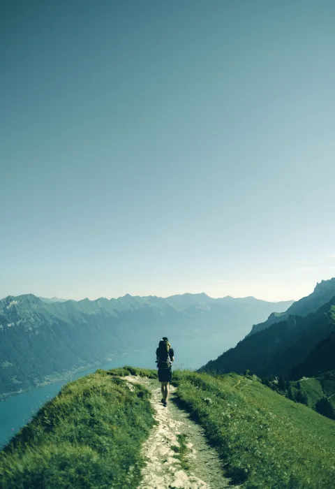 Person mit Rucksack an einem Aussichtspunkt, Blick über eine Bergkette und einen See im Dunst